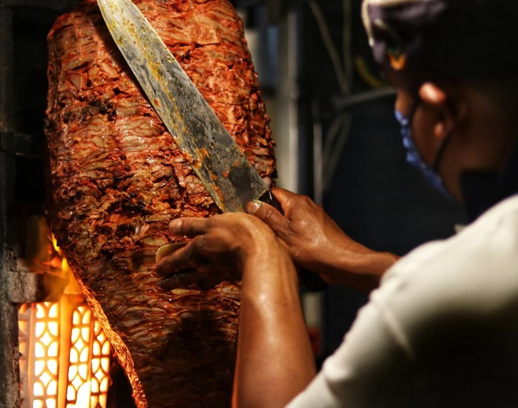 Un couteau racle une broche verticale : la viande tombe en lamelles, la braise éclaire le geste.