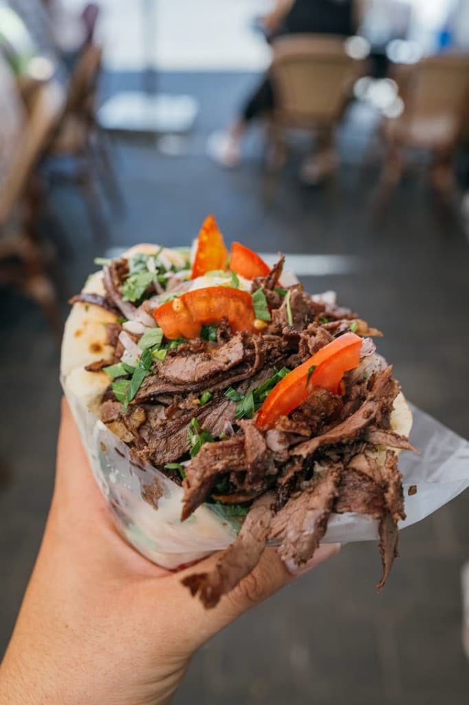 Un kebab tenu à la main, pain ouvert, viande, tomate, oignon, sauce blanche, papier qui dépasse.